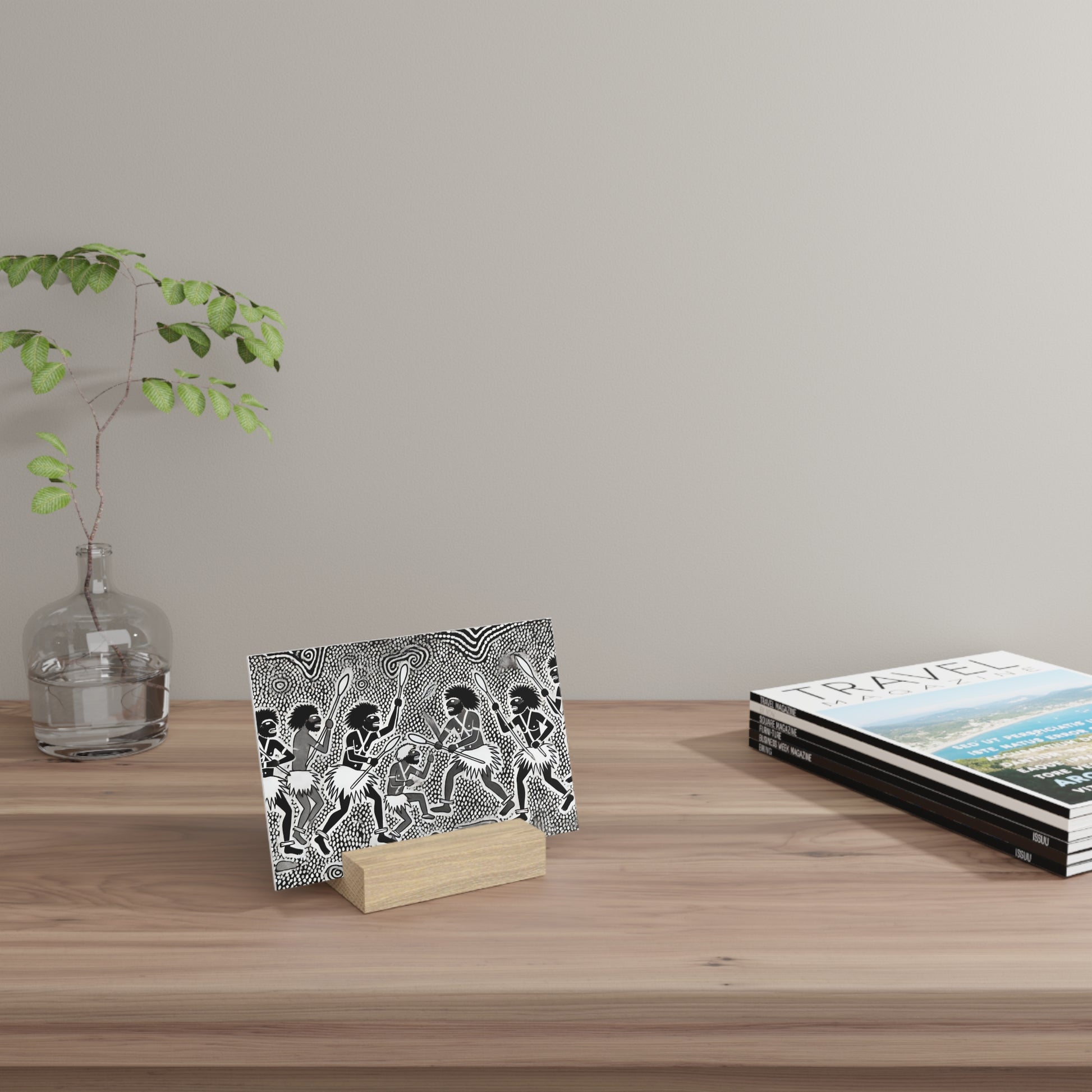 Clear glass vase with a green leafy branch, a black-and-white illustrated card on a wooden stand, and a travel magazine stack on a wood