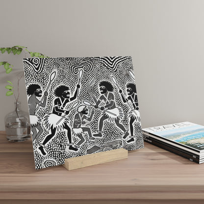Black-and-white canvas print of tribal dancers with spears, propped on a wooden block on a light wood table beside magazines.
