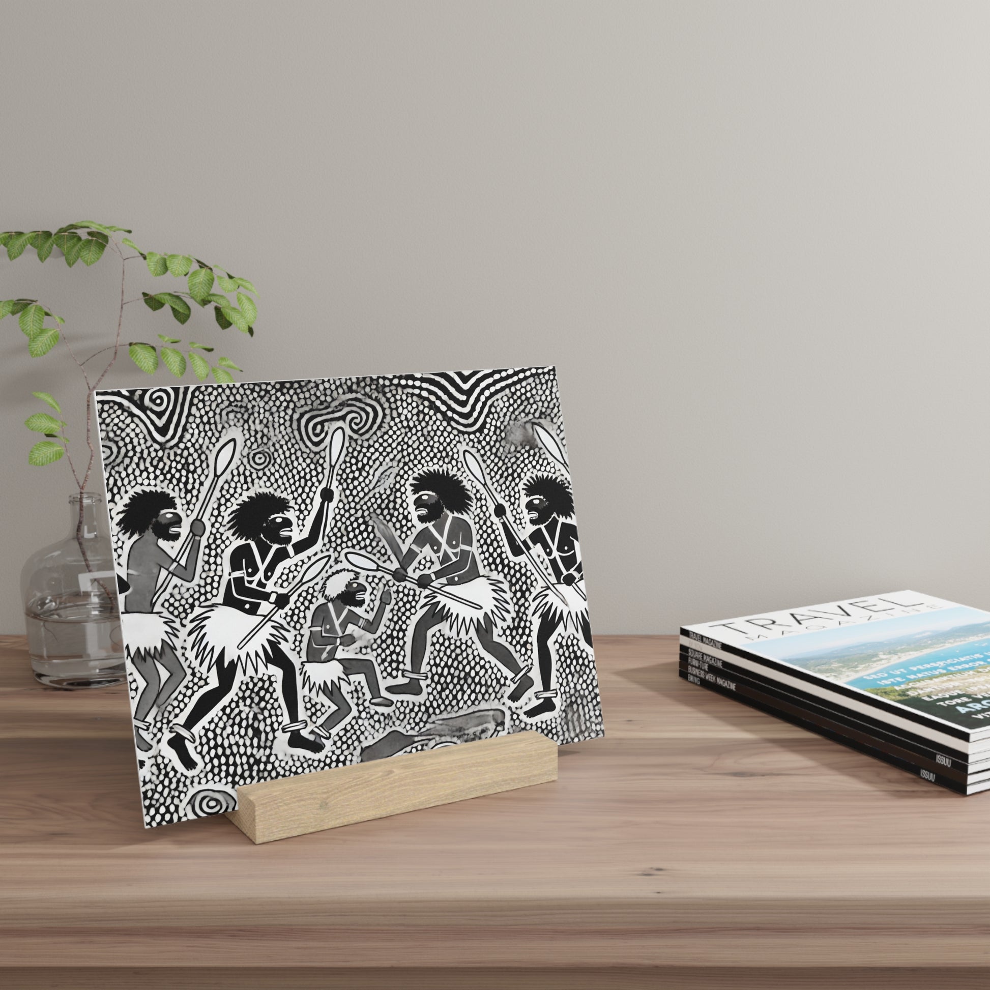 Black-and-white tribal print on a rectangular panel rests on a light wooden block on a desk, with a glass vase of greenery to the left.