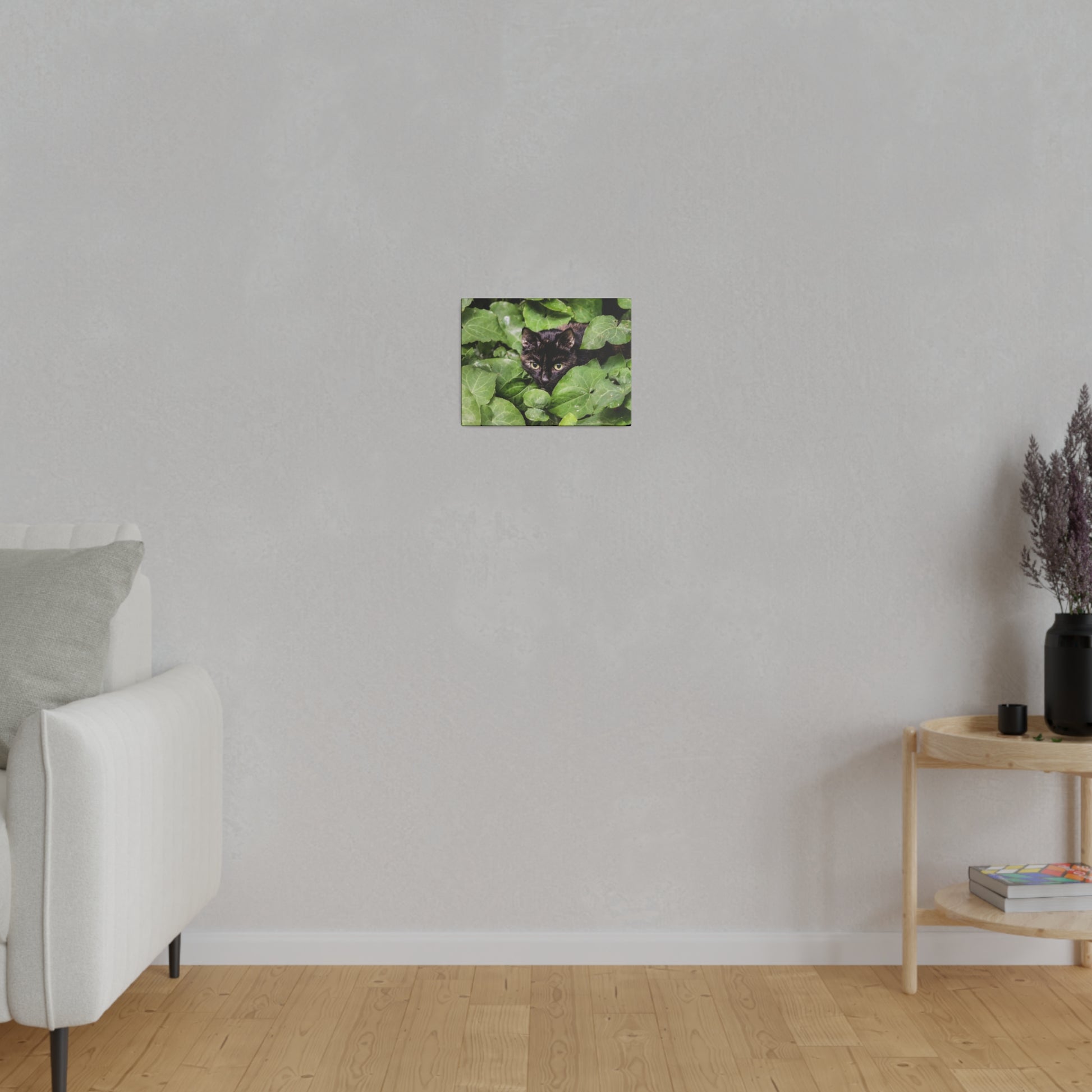 Small square photo of a black cat peeking from green leaves, hung on a light gray wall.