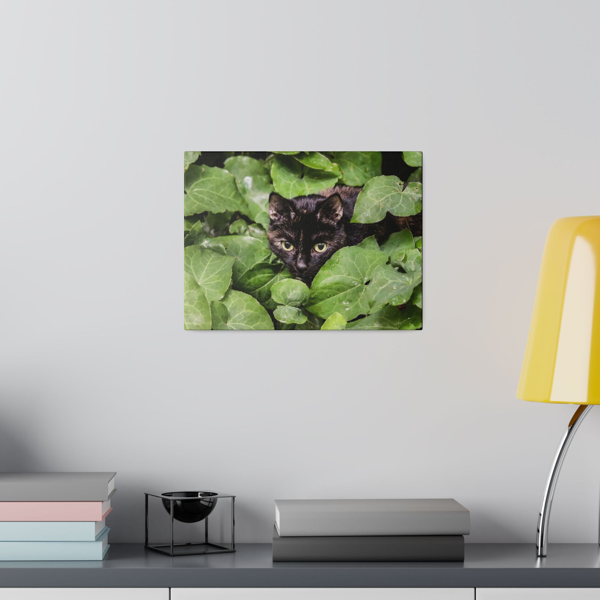 Poster-style photo of a black cat peeking through green ivy leaves on a white wall.