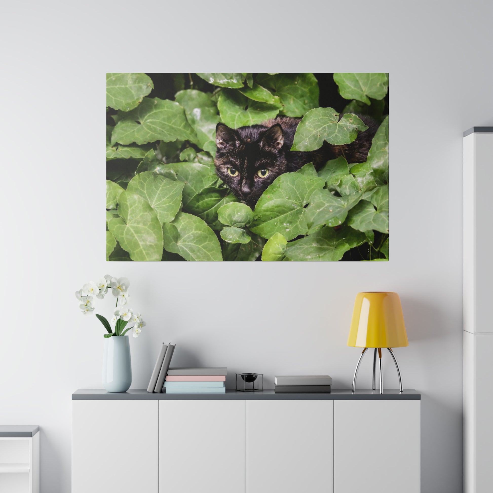 Black cat with yellow-green eyes peeking through dense green leaves mounted on a wall above a cabinet.