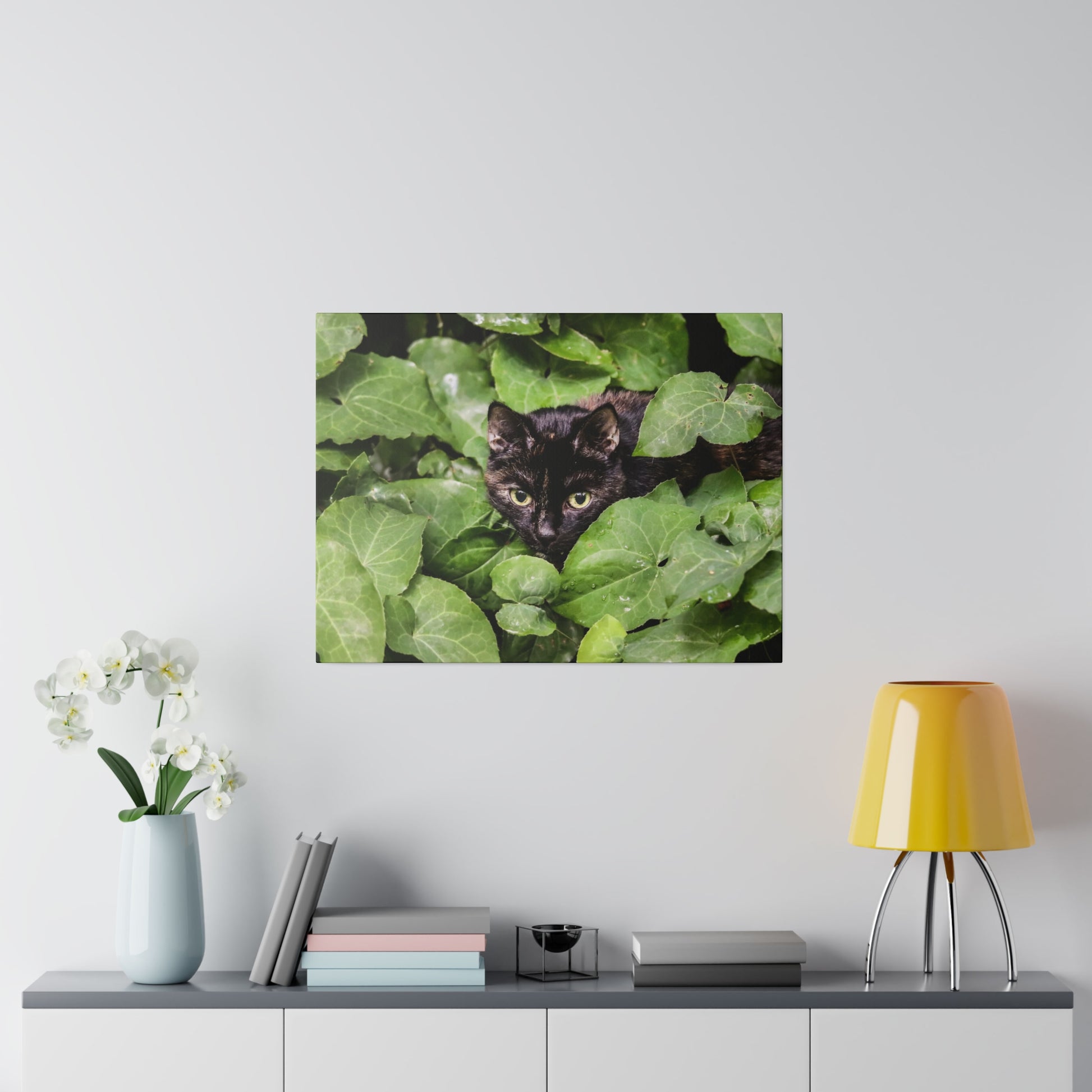 Framed print of a black cat peeking through dense green ivy leaves.