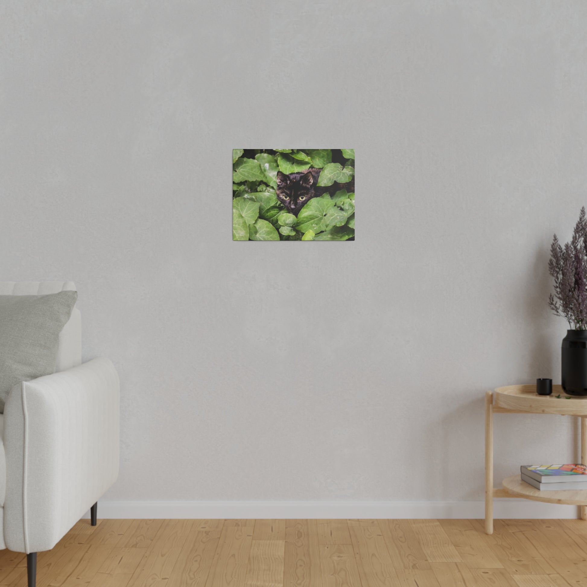 Small square wall art on a gray wall, showing a black cat peeking through green leaves.