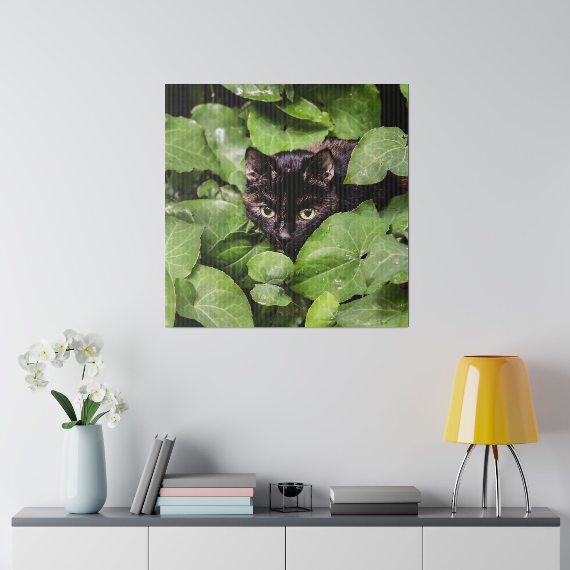 Square wall art depicting a black cat with green eyes peeking through dense green leaves.