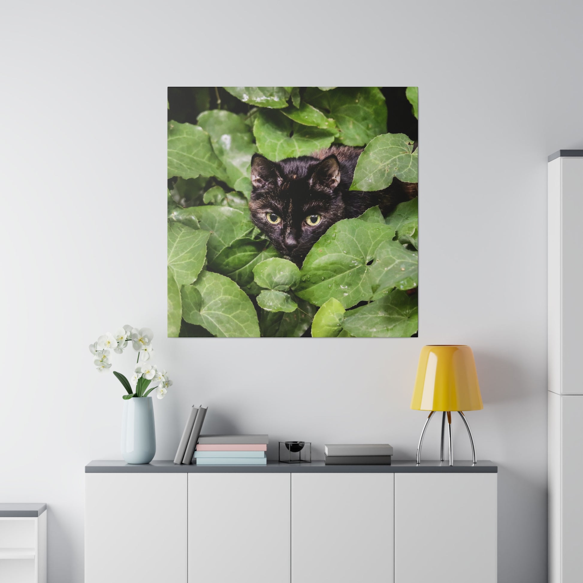 Framed wall art of a black cat peeking through green leaves, hung above a white cabinet with a yellow lampshade and vase.