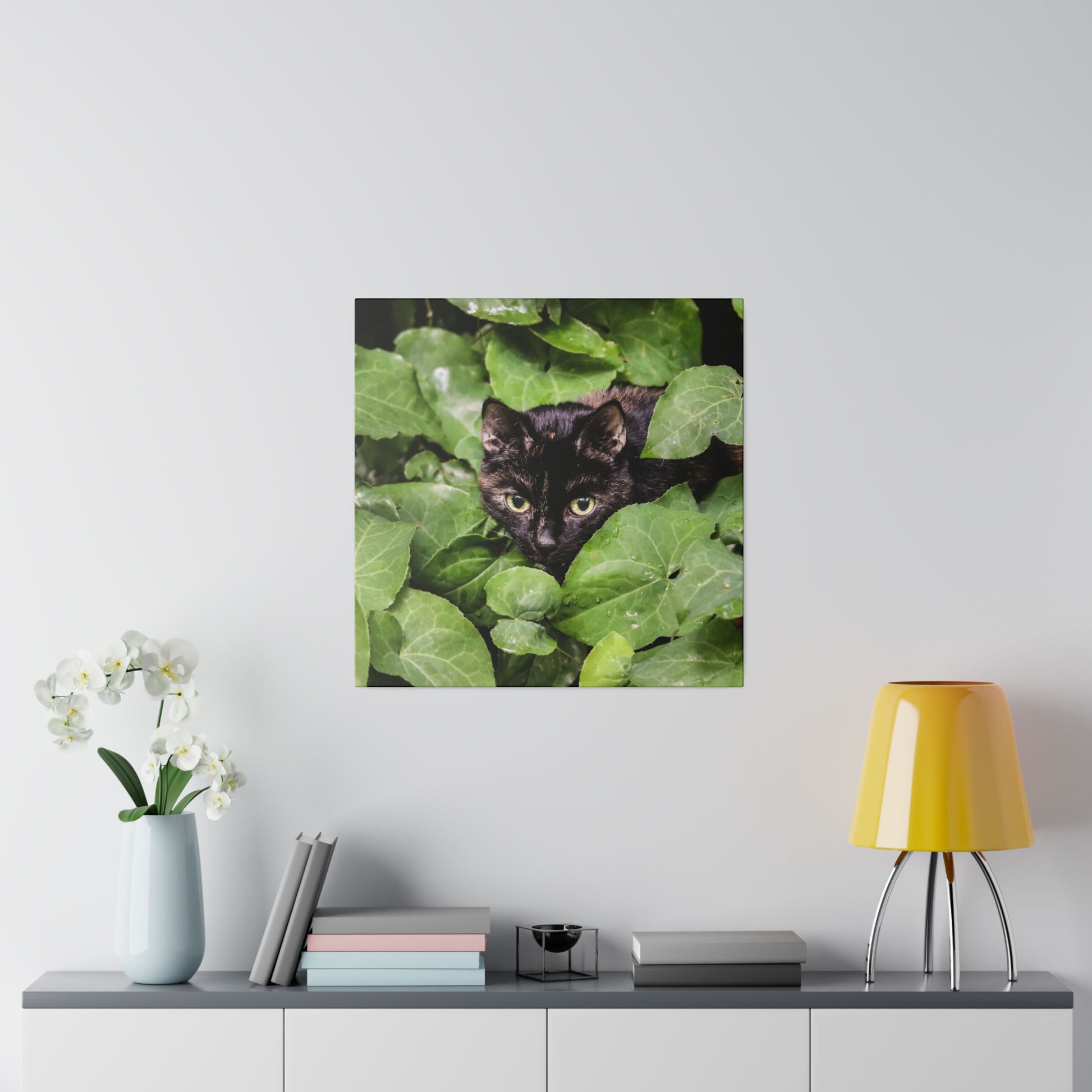 Square photo of a black cat peeking through dense green leaves.