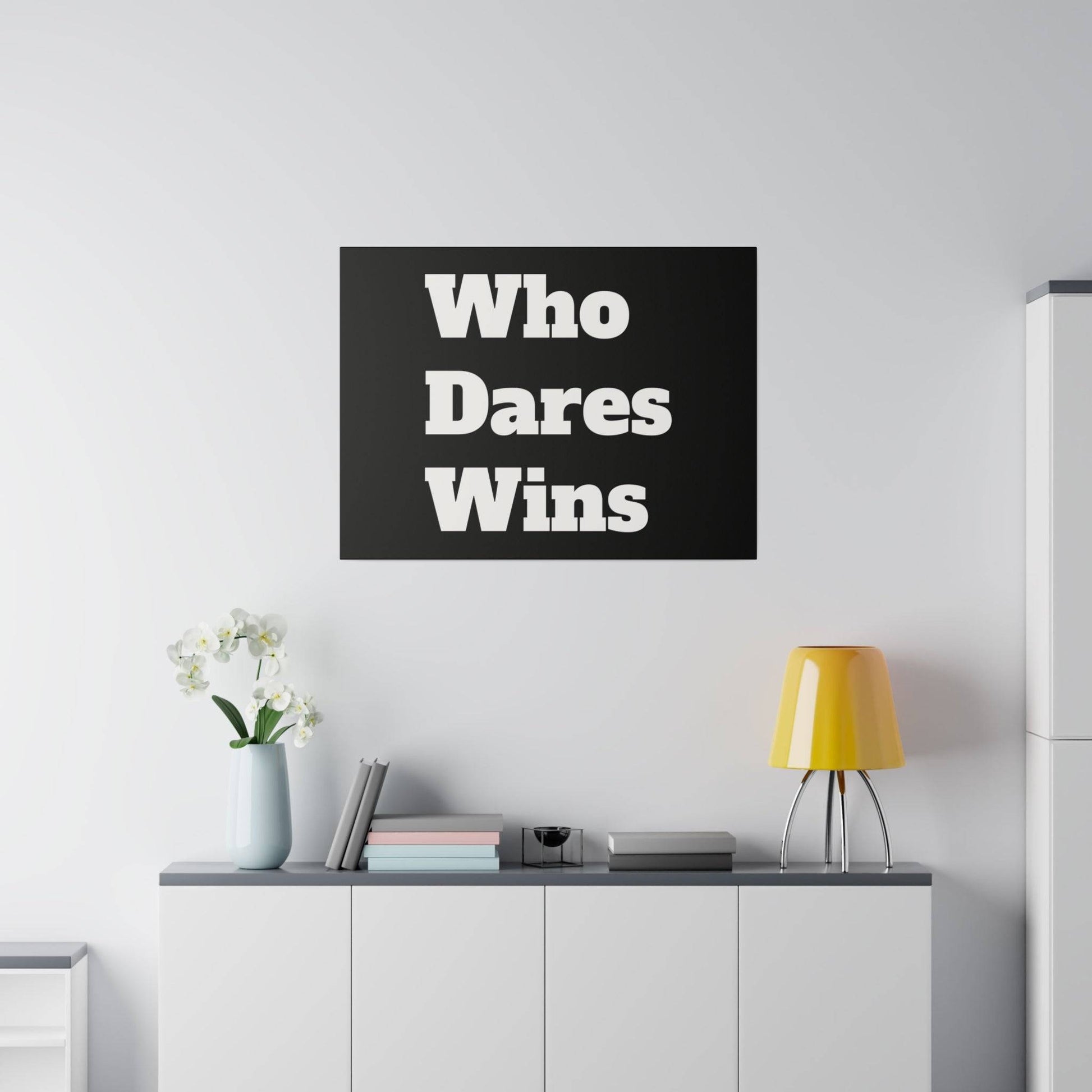 Black rectangular wall art with bold white text "Who Dares Wins" above a white cabinet, beside a vase of white flowers and a yellow lamp.