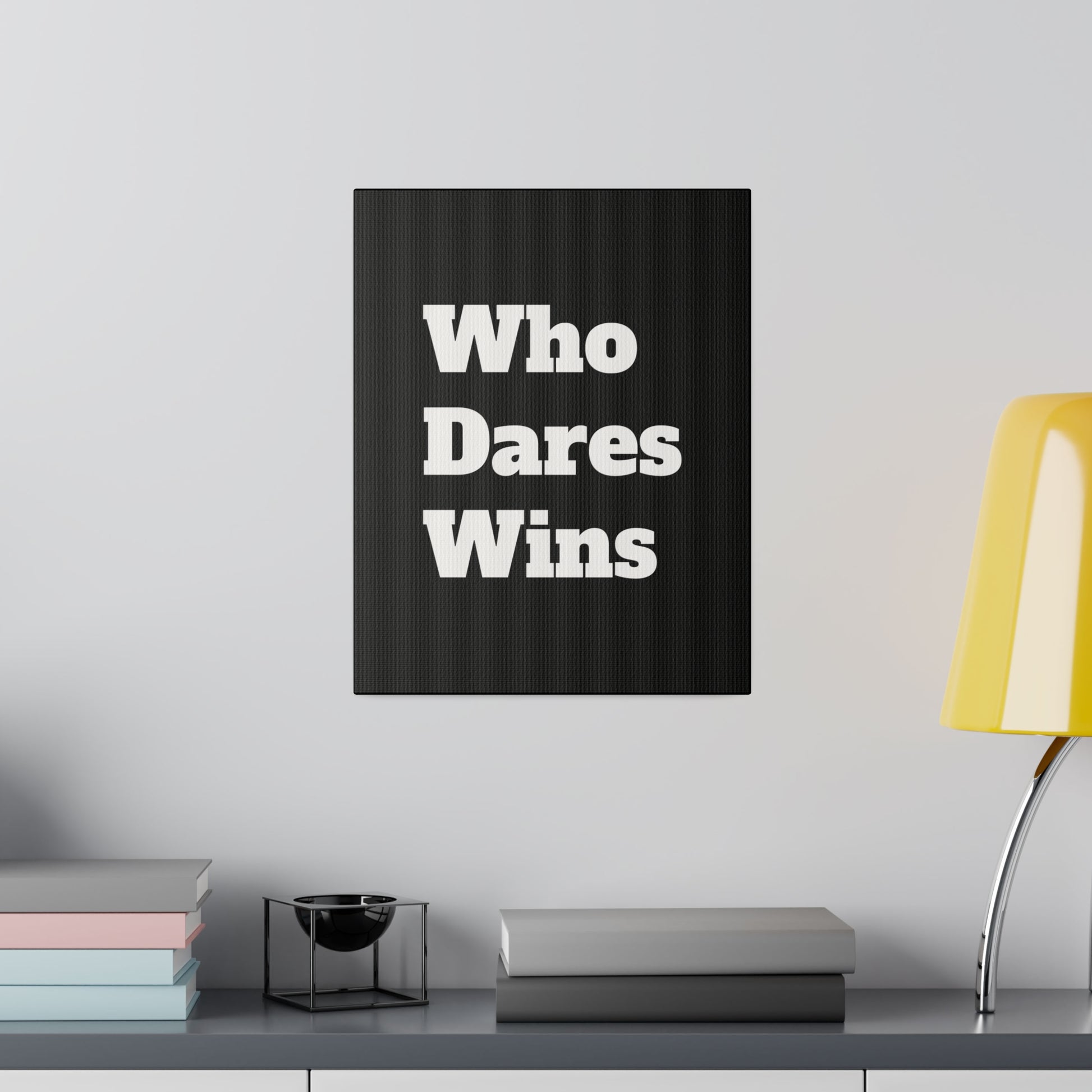 Black canvas with bold white text "Who Dares Wins" mounted on a light gray wall above a shelf with books and a yellow desk lamp.