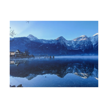 Snow-capped mountains loom over a calm lake with a wooden dock and houses on the left, reflecting in the water.