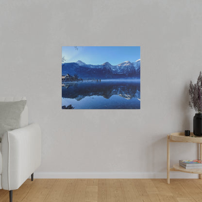 Square wall art showing a blue alpine lake with snow-capped mountains and a village, reflected in the calm water.